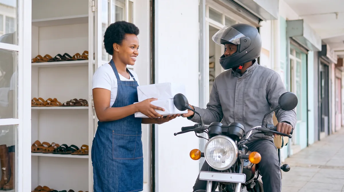 Nairobi leather sandal maker smiling as she hands wrapped parcels to a delivery motorbike rider outside her workshop